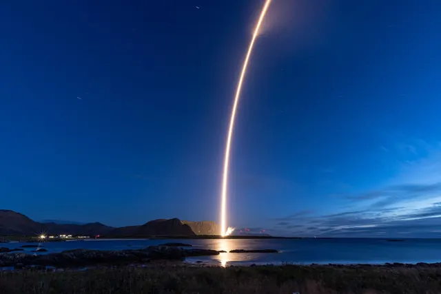 A rocket’s glowing trail arcing up from Andøya in blue twilight, over water and mountains