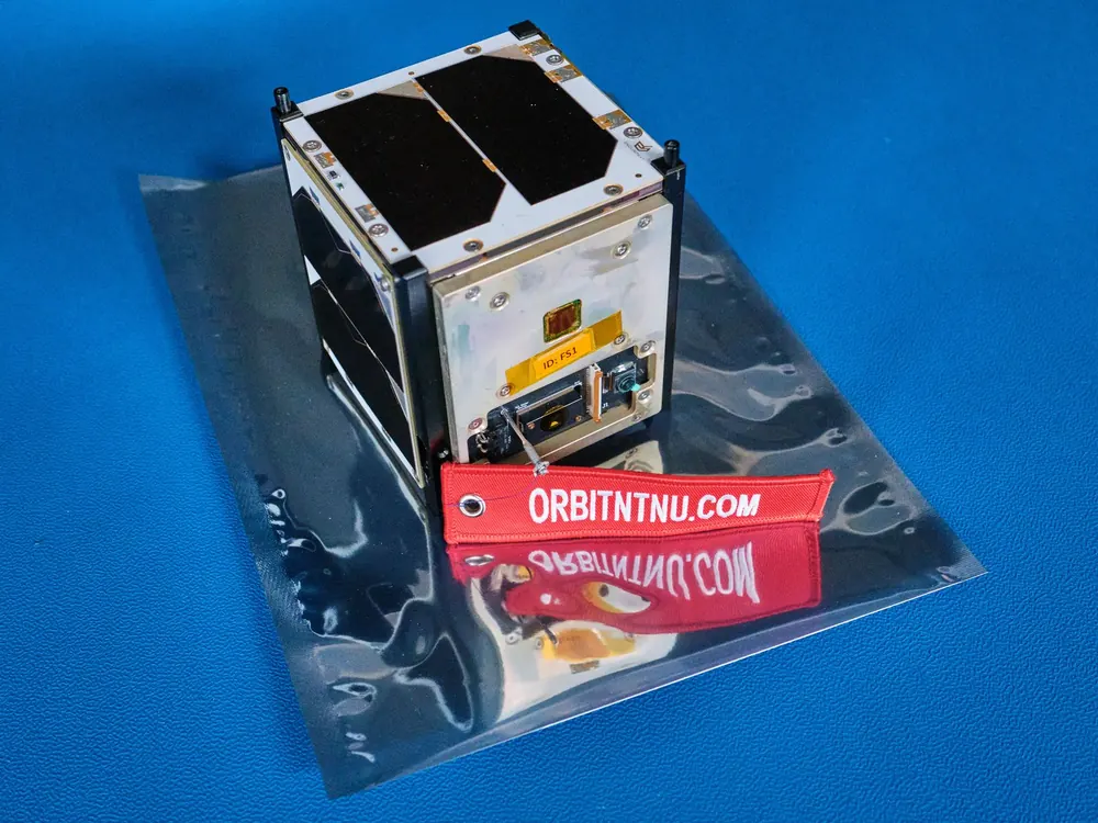 FramSat-1, a small cube-shaped satellite with solar panels, on an anti-static bag with red Orbit NTNU tags