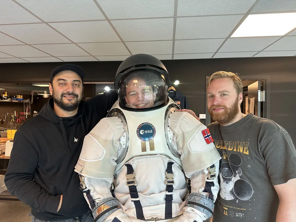 Freider in a white spacesuit and helmet, between two people from Nåva Space